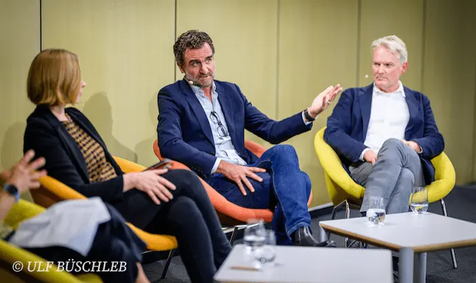 Eine Frau und zwei Männer sitzen nebeneinander auf einer Bühne. Der Mann in der Mitte spricht.
