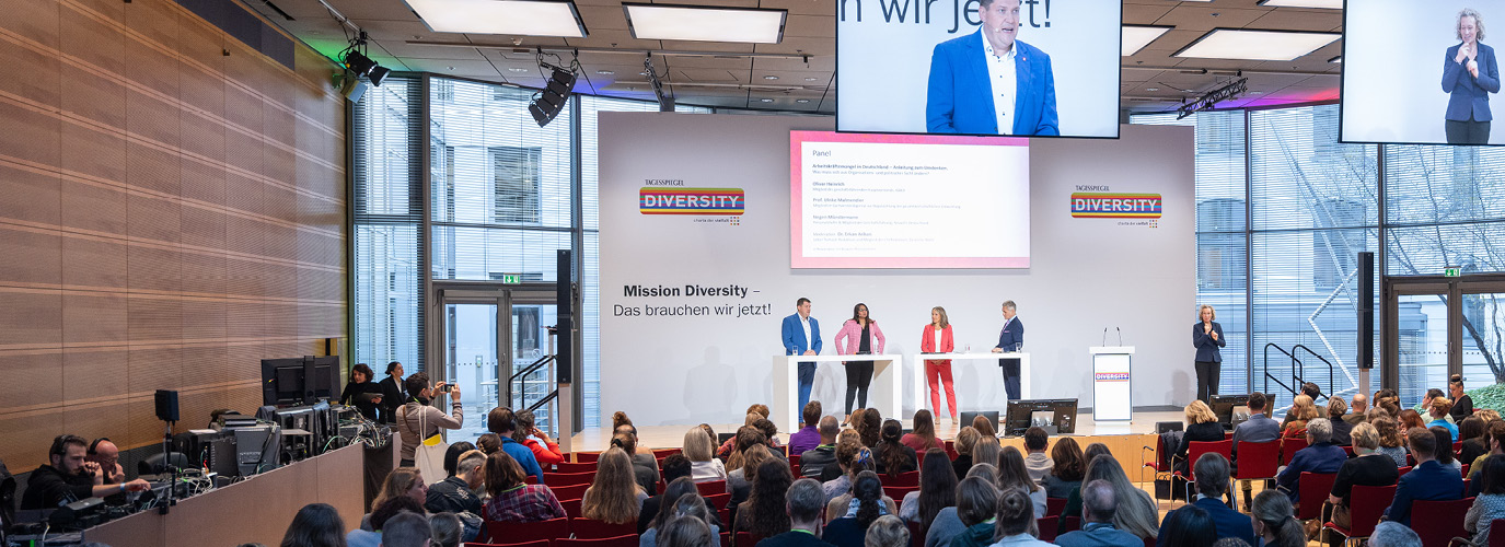 Das Foto zeigt einen Ausschnitt der Konferenz „DIVERSITY 2023“. Mehrere Menschen stehen auf einer Bühne, davor sitzt ein großes Publikum