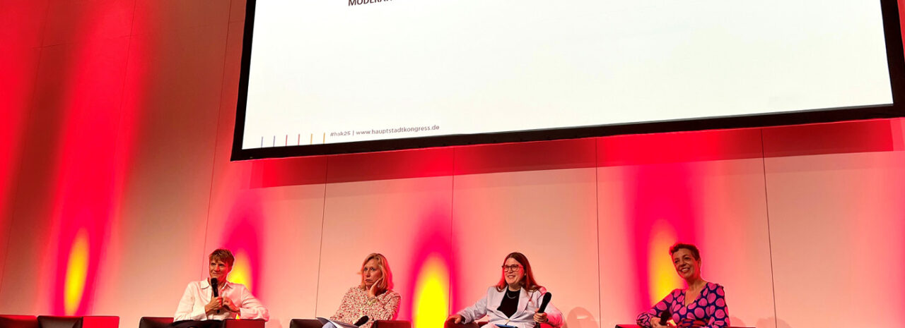 Vier Frauen sitzen auf einer Bühne in Sesseln und nehmen unter roter und gelber Beleuchtung an einer Podiumsdiskussion teil, während über ihnen ein großer leerer Bildschirm angezeigt wird.