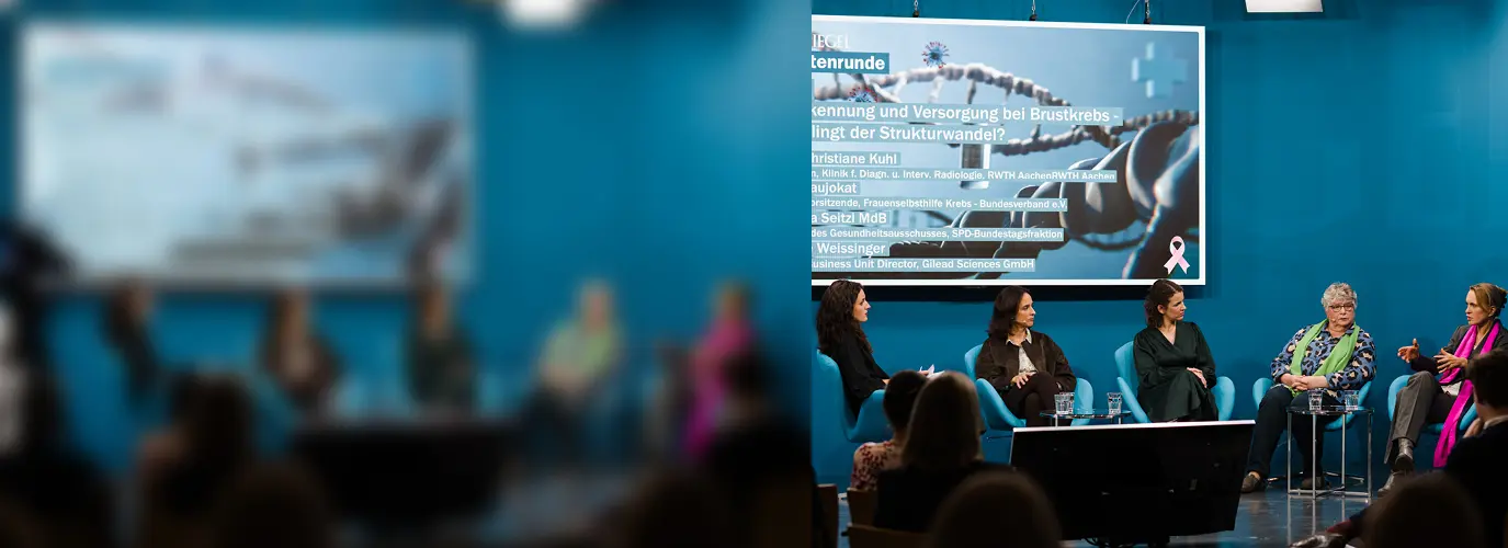 Eine Gruppe von fünf Frauen sitzt auf der Bühne vor einem blauen Hintergrund und einem großen Bildschirm und führt eine Diskussion. Die linke Seite des Bildes ist unscharf, während die rechte Seite scharf abgebildet ist. Eine Gruppe von fünf Frauen sitzt auf der Bühne vor einem blauen Hintergrund und einem großen Bildschirm und führt eine Diskussion. Die linke Seite des Bildes ist unscharf, während die rechte Seite scharf abgebildet ist.