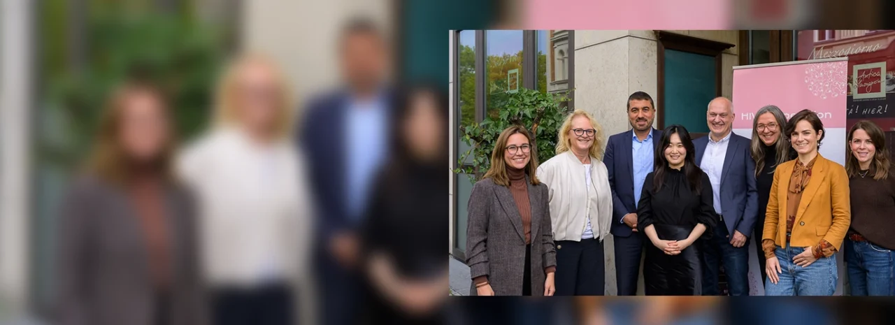 Eine Gruppe von acht Personen, fünf Frauen und drei Männer, steht im Freien zusammen und lächelt in die Kamera. Die Personen sind leger gekleidet und scheinen an einer geschäftlichen Veranstaltung teilzunehmen.