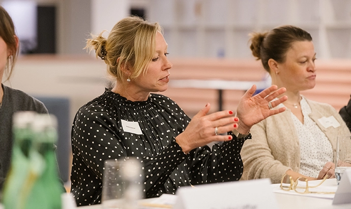 Eine Frau in einer schwarzen gepunkteten Bluse gestikuliert mit ihren Händen, während sie in einer Besprechung an einem Tisch mit Getränken und Notizbüchern sitzt. Eine andere Frau hört neben ihr zu.