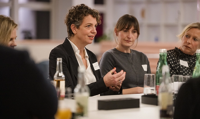 Vier Frauen sitzen an einem Tisch und diskutieren, vor ihnen stehen Getränke und Flaschen. Eine Frau in einer Anzugsjacke gestikuliert, während sie spricht, und die anderen hören aufmerksam zu. Es scheint sich um eine berufliche Besprechung zu handeln.