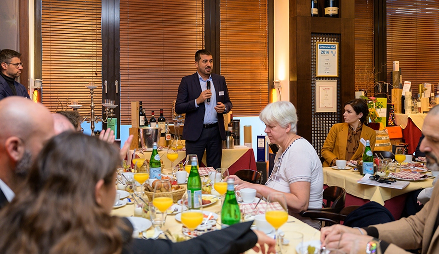 Ein Mann steht und spricht mit einem Mikrofon zu einer Gruppe von Menschen, die in einem gut beleuchteten Innenraum an Tischen mit Essen, Orangensaft und Wasserflaschen sitzen.